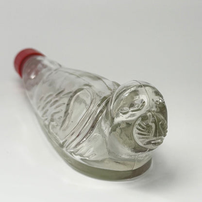 Vintage tysk figurflaske i klart formpresset glas, udformet som en liggende sæl – Seehund.

Flasken har detaljeret relief med sælens hoved, luffer og krop. Hoved og snude er udført i massivt glas.

Denne type figurflaske blev fremstillet i Tyskland som emballage til spiritus. Tilsvarende eksemplarer ses med originale etiketter mærket bl.a. “Seehund” og “Deutsches Erzeugnis”. Producenten af dette eksemplar er ikke identificeret med sikkerhed.

Med original/lignende ældre rød skrueprop i plast med kork på ind