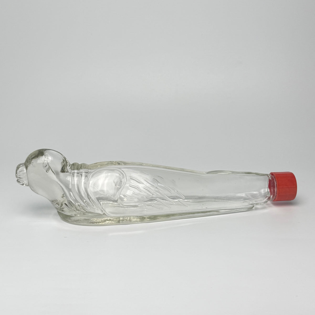 Vintage tysk figurflaske i klart formpresset glas, udformet som en liggende sæl – Seehund.

Flasken har detaljeret relief med sælens hoved, luffer og krop. Hoved og snude er udført i massivt glas.

Denne type figurflaske blev fremstillet i Tyskland som emballage til spiritus. Tilsvarende eksemplarer ses med originale etiketter mærket bl.a. “Seehund” og “Deutsches Erzeugnis”. Producenten af dette eksemplar er ikke identificeret med sikkerhed.

Med original/lignende ældre rød skrueprop i plast med kork på ind